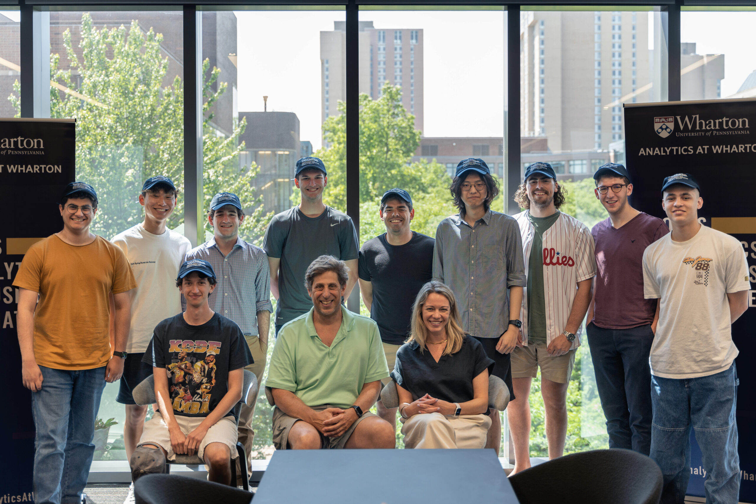 A group of people, some sitting and some standing, posed indoors with a backdrop of large windows showing an urban landscape. They wear casual clothing, some with matching caps. Two banners are visible, indicating affiliation with Wharton School, University of Pennsylvania.