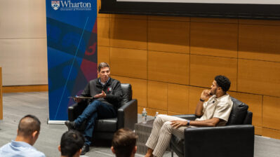 A discussion event featuring two individuals sitting in armchairs on a stage. One holds a tablet and gestures while speaking. A Wharton University of Pennsylvania banner is visible in the background. A small group of people listens in the foreground.