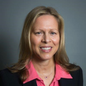 Headshot of a smiling individual with long hair wearing a pink shirt and dark blazer against a plain background.