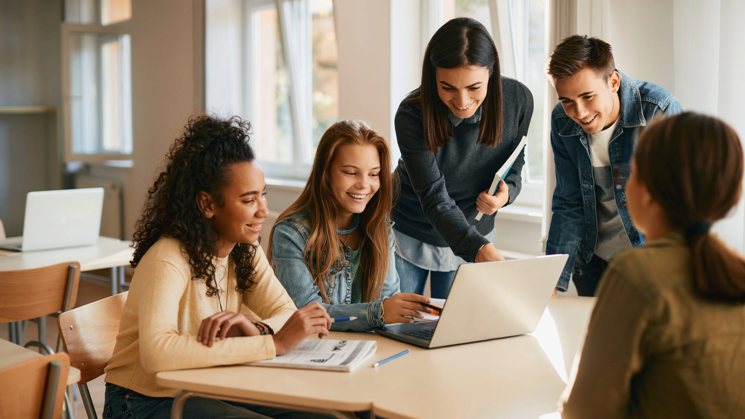 A group of young adults gathered around a laptop in a classroom setting, engaged in a collaborative activity or study session, smiling and interacting.