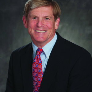 A headshot of a person wearing a suit, blue shirt, and patterned red tie, smiling against a dark background.