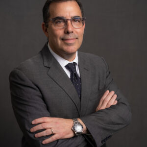 A headshot of a person wearing a suit and tie with crossed arms, glasses, and a watch, set against a plain background.