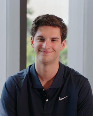 Headshot of a person smiling, wearing a navy blue Nike polo shirt, with a window and greenery in the background.