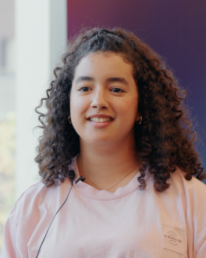 A person with curly hair and wearing a pink t-shirt is smiling. They have a microphone attached to their shirt. The background is a gradient of purple and a window offering natural light.