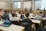 A university lecture hall with a group of students seated at desks, laptops open. Two presenters stand in front, near projected screens displaying a presentation.