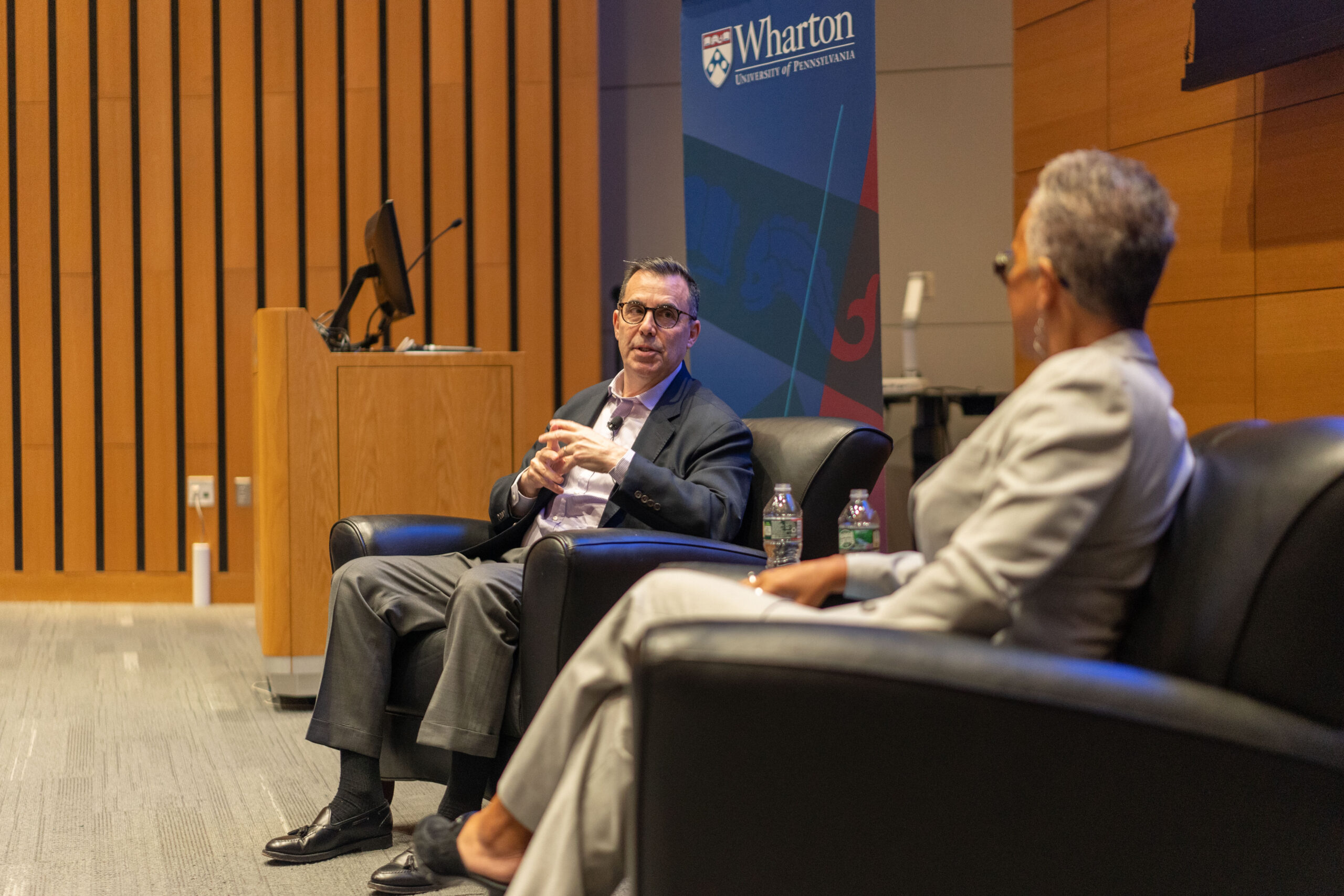 A discussion between two individuals seated in armchairs in an academic setting, with a Wharton School of the University of Pennsylvania banner in the background.