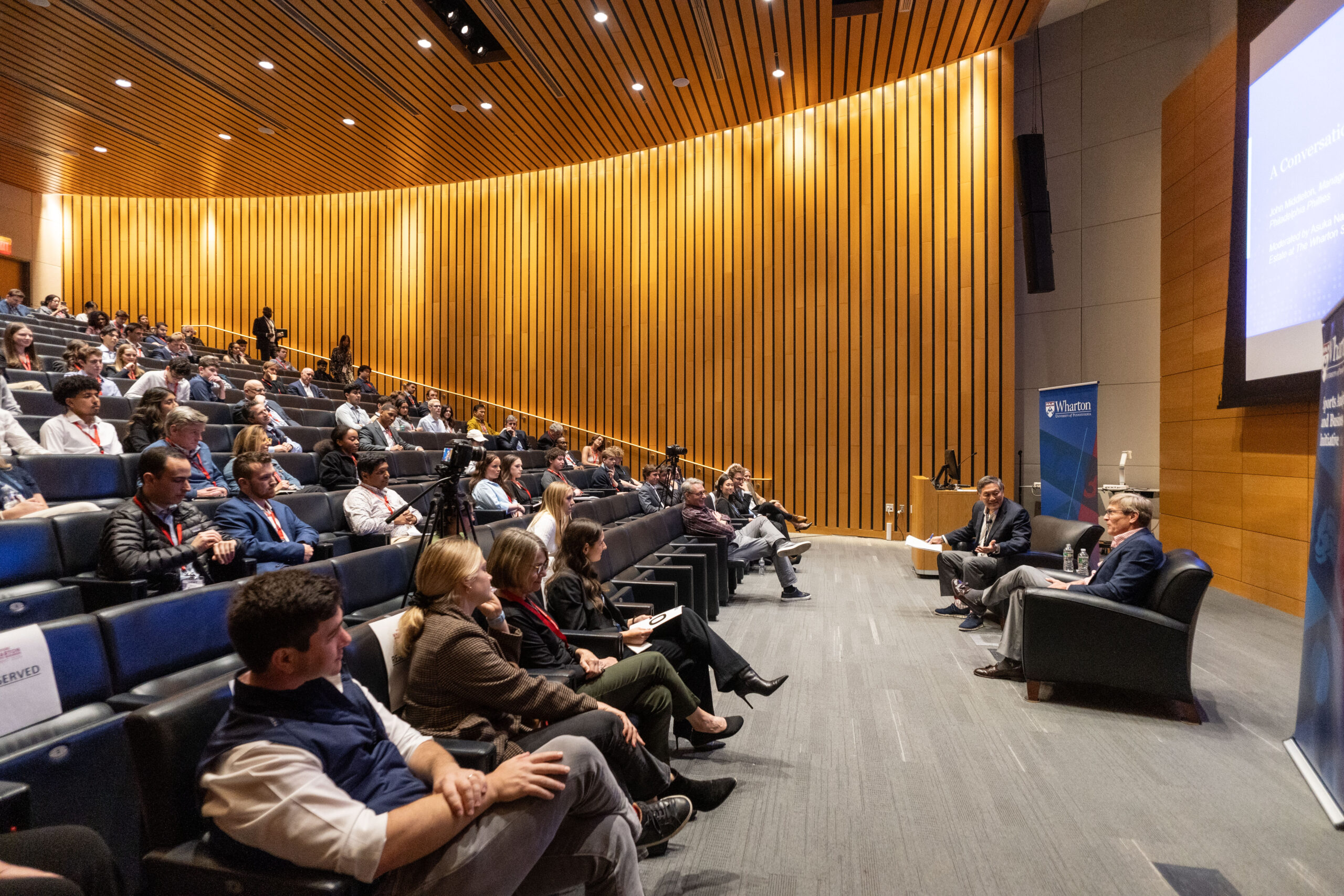 An auditorium with people sitting and listening to two speakers engaged in a discussion. The room features a wooden panel design and a presentation screen displaying an event title.