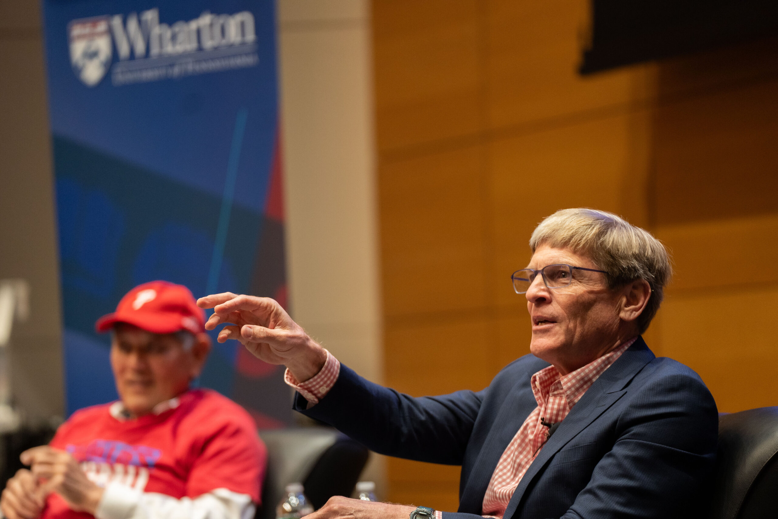 A person speaking and gesturing while seated in a panel discussion, with a Wharton School banner in the background. Another person, wearing a red cap and shirt, is also seated nearby.