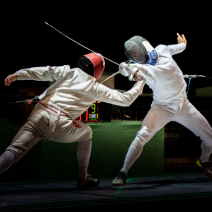 Two fencers in protective gear and helmets are engaged in a duel on a fencing piste.