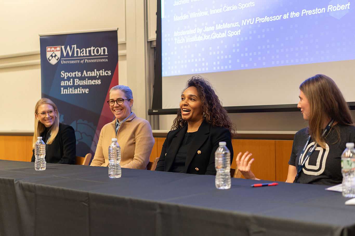 2024 WSBS A panel of four people sits at a long table with water bottles. They are engaged in a discussion, likely related to the Wharton Sports Analytics and Business Initiative, as indicated by the banner in the background. They appear to be in a lecture or