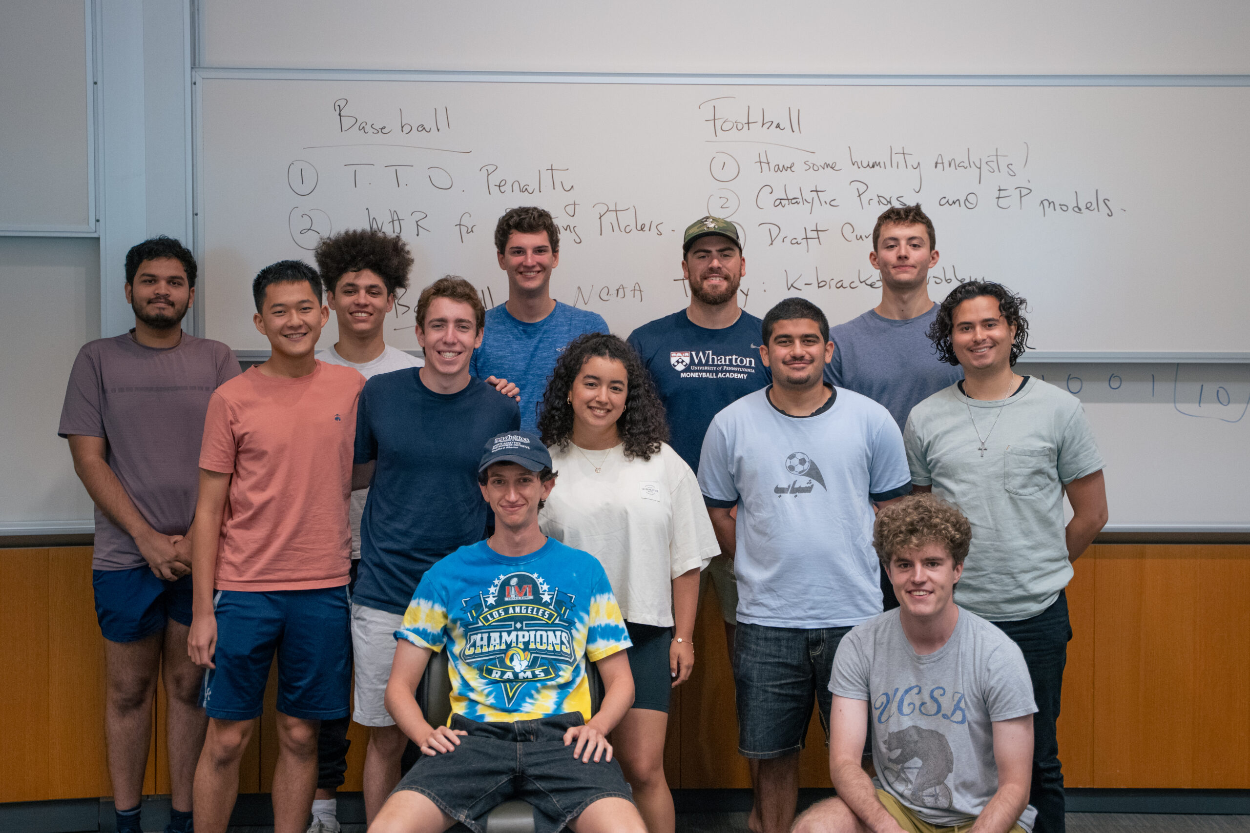 Brill's Cohort A group of people smiling in front of a whiteboard with notes about baseball and football.