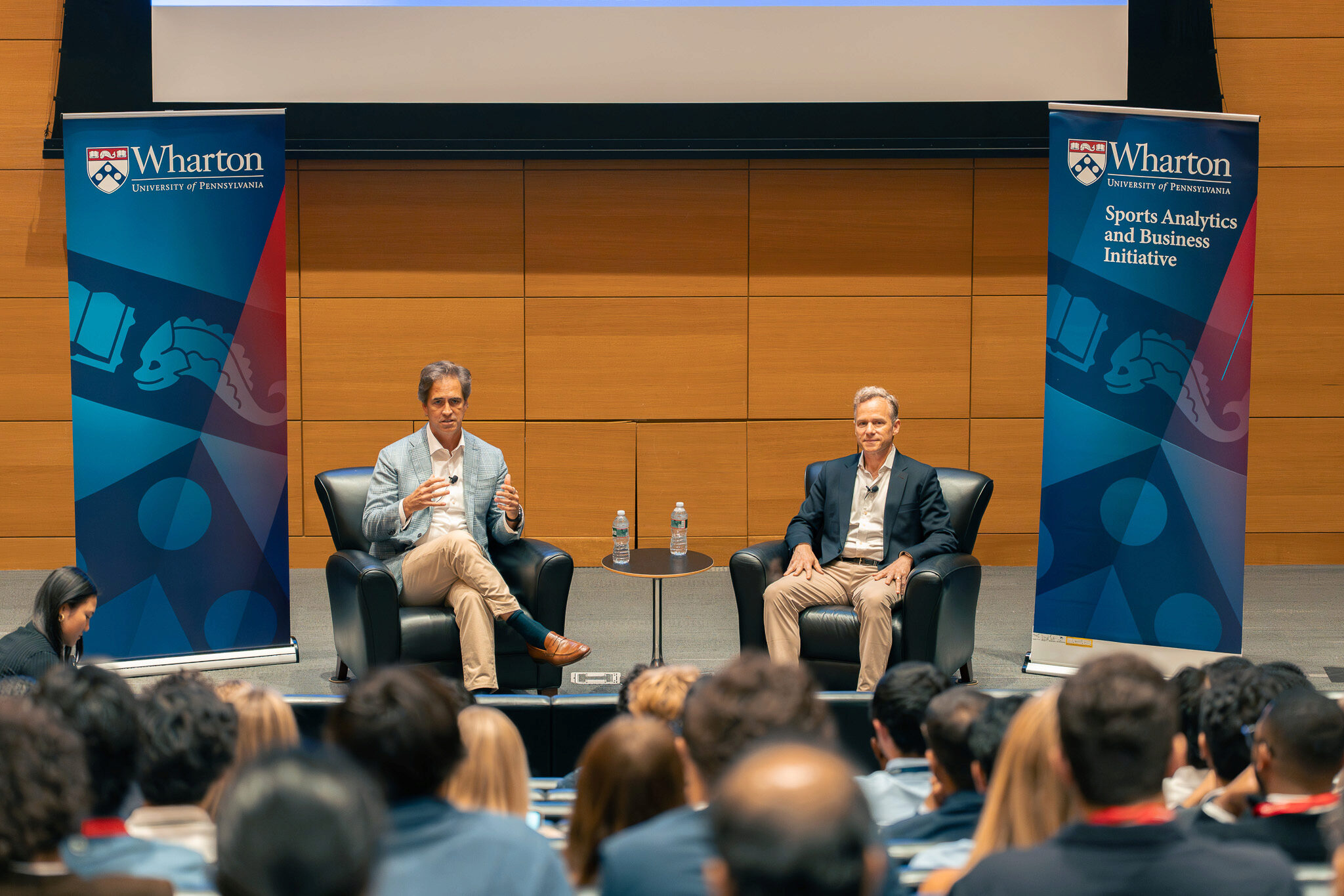 DSC00531 Two people seated on stage at a Wharton event, discussing sports analytics and business. Audience members are visible in the foreground.