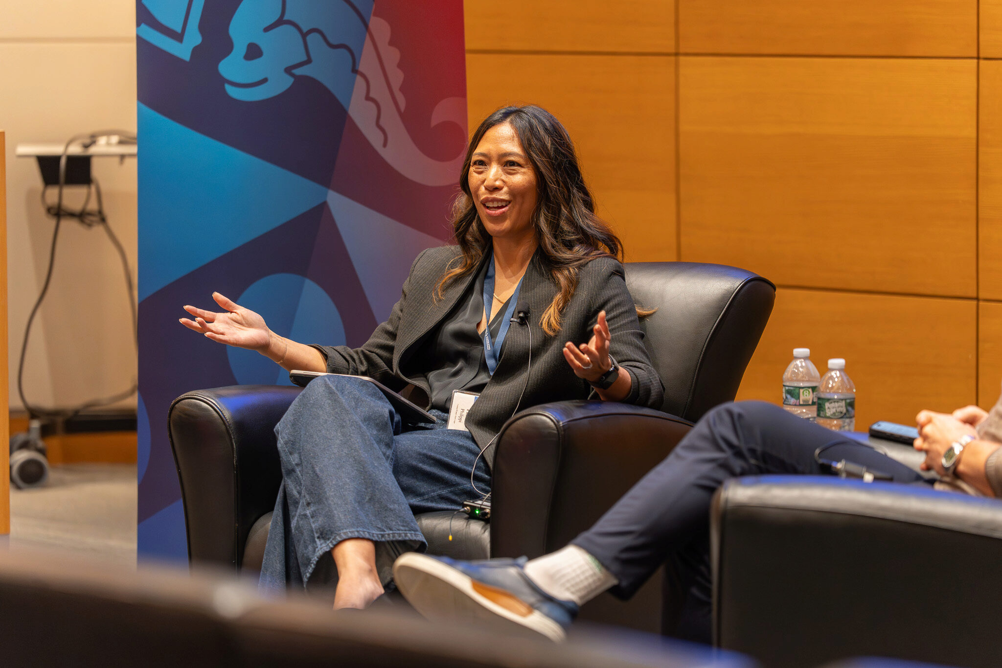 DSC00769 A person is speaking while seated in a black chair, gesturing with both hands. A colorful banner and water bottles are visible nearby.