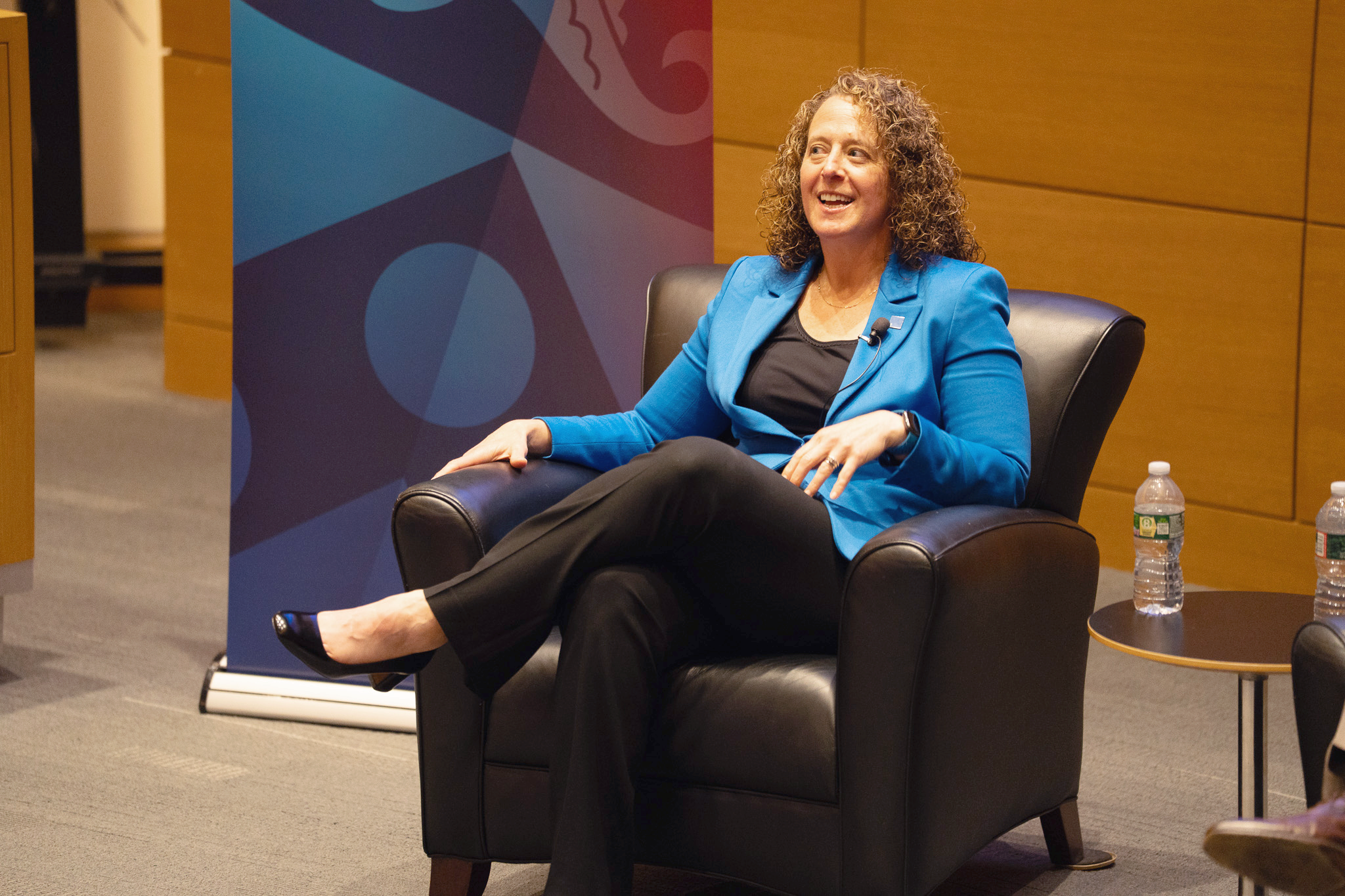 Gelman Summit A person in a blue blazer sits in a black armchair, smiling during a panel discussion. Bottles of water are on a table nearby.