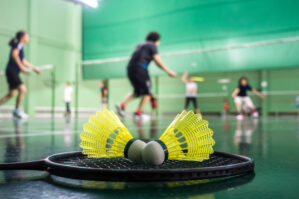 Close-up of a badminton racquet and shuttlecocks on a court with several people playing in the background.
